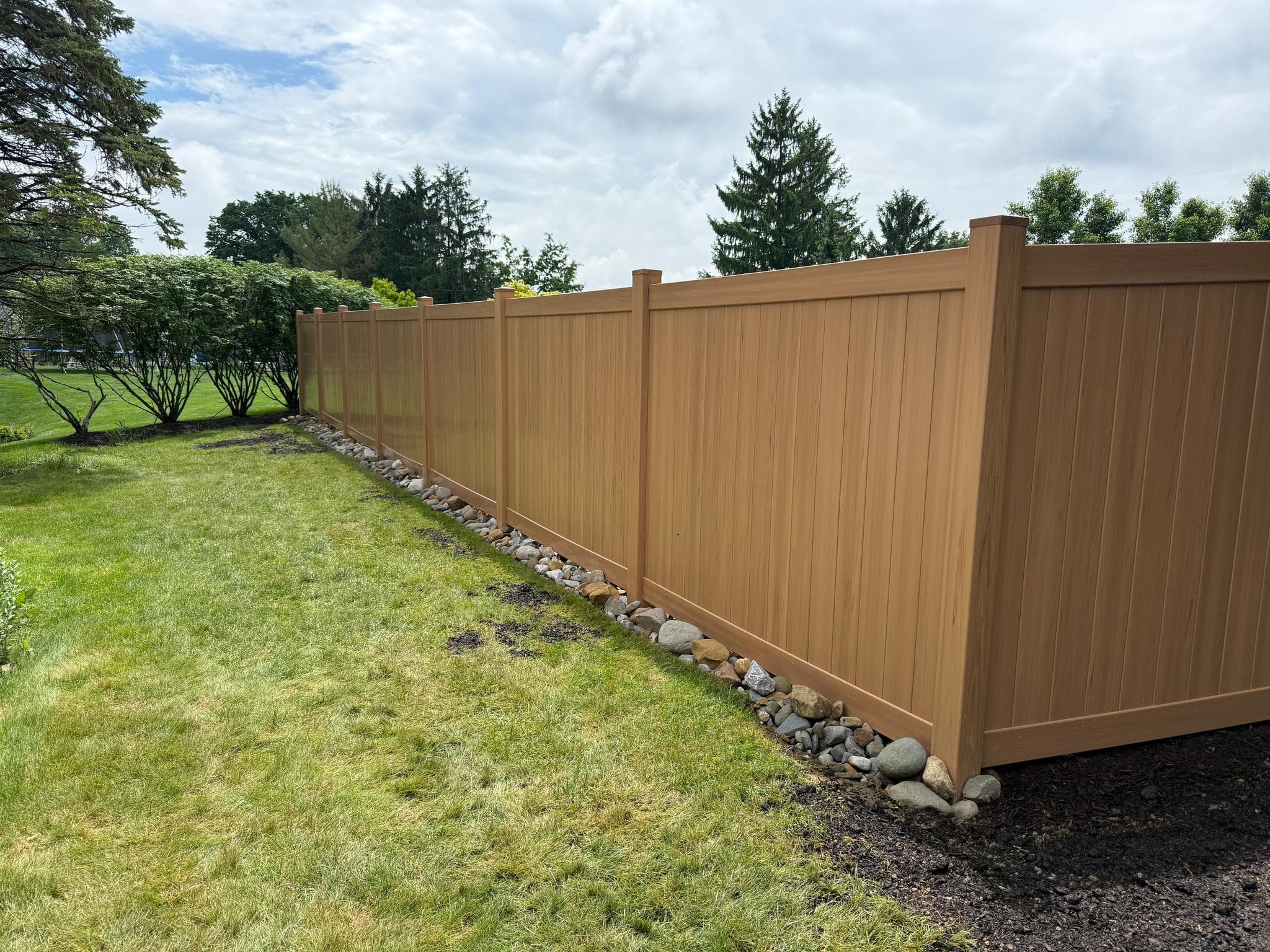 Cedar-tone vinyl privacy fence installation with a river-rock border in a Pittsburgh backyard